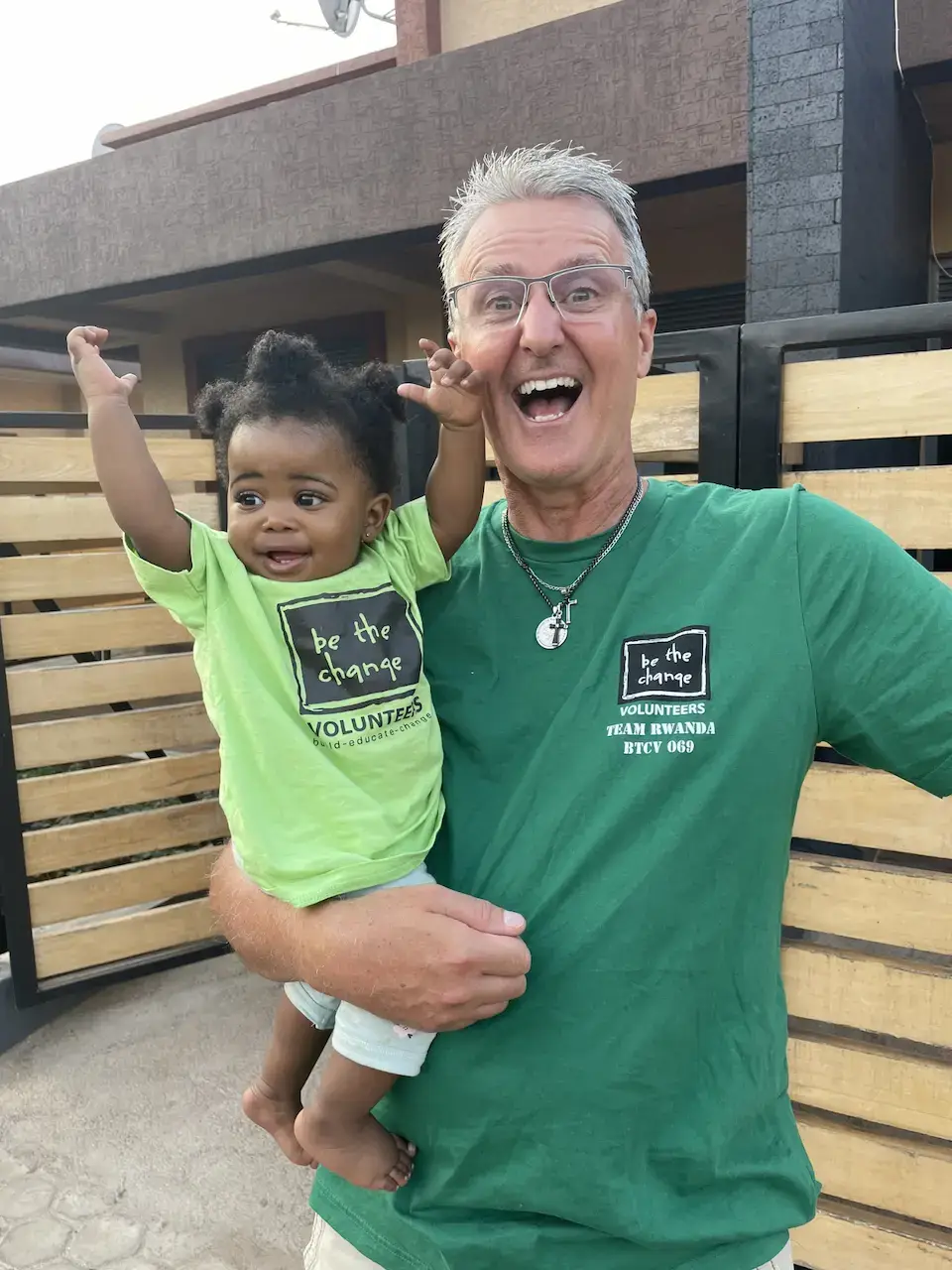 A man wearing a green "Be the Change Volunteers Team Rwanda" shirt is holding a young child who is also wearing a green "Be the Change Volunteers" shirt.