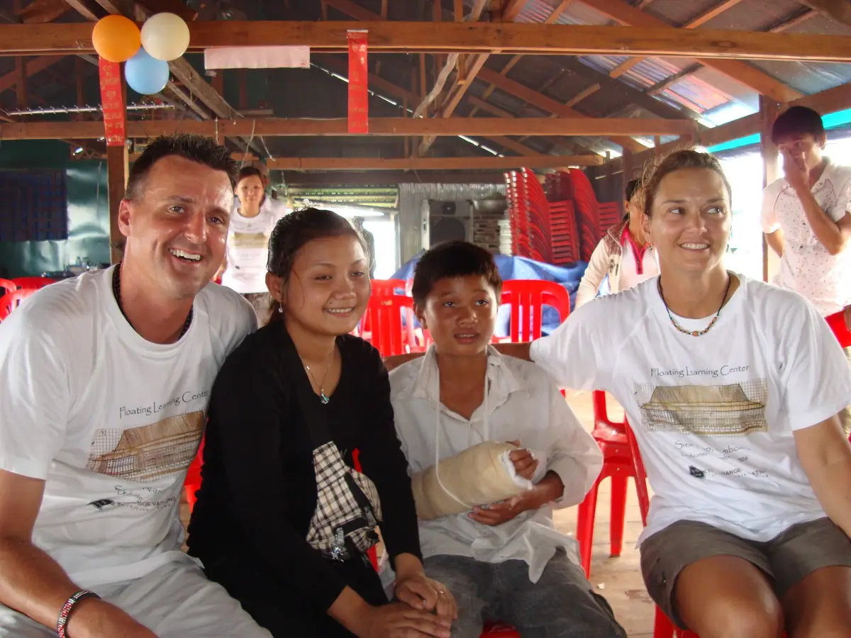 Two volunteers in "Floating Learning Center" T-shirts sit smiling beside a young woman and a boy with his arm in a cast, under a wooden roof hung with balloons in Cambodia.