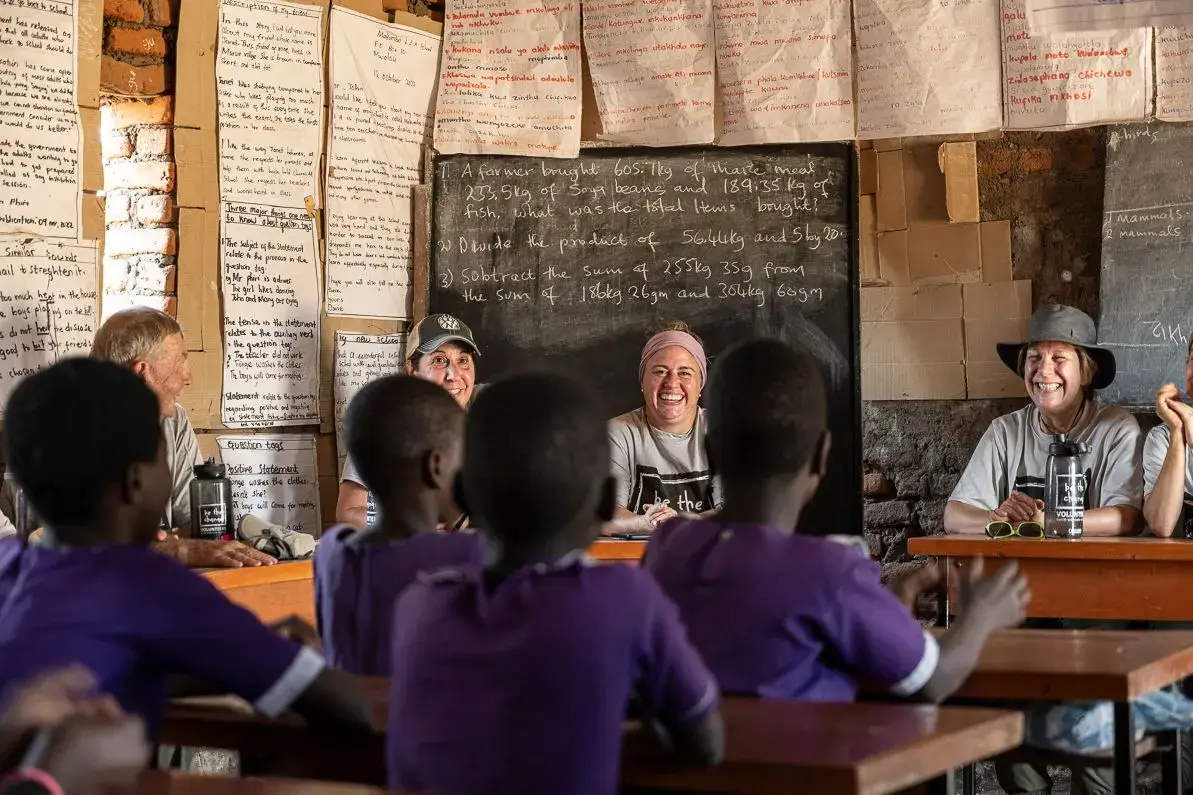 a classroom with a chalkboard showing math problems, where a group of adults and children are sitting at desks facing each other