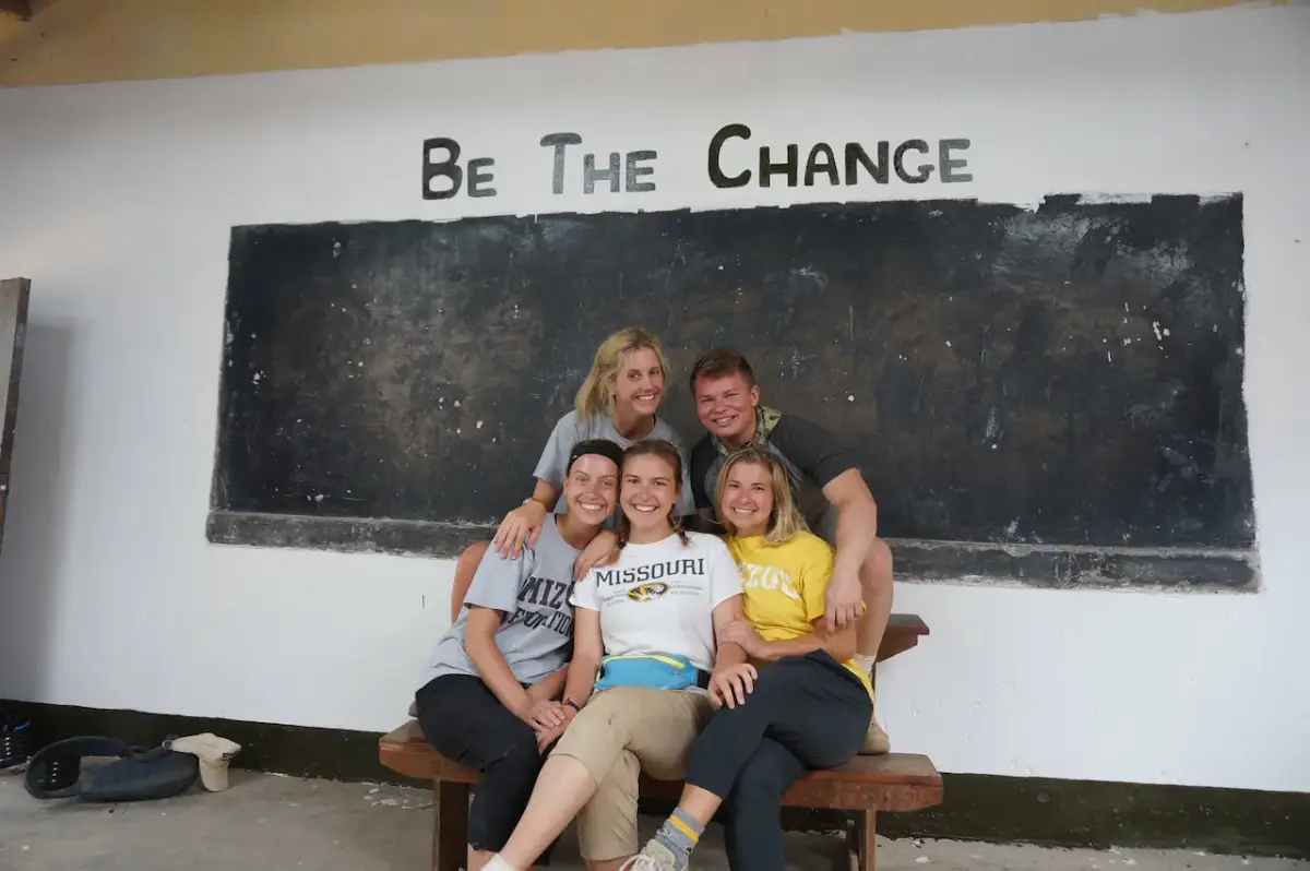 A group of volunteers, five young adults—three women in the front row and a woman and a man in the back row—are sitting and smiling in front of a chalkboard with the text "BE THE CHANGE" written above it.