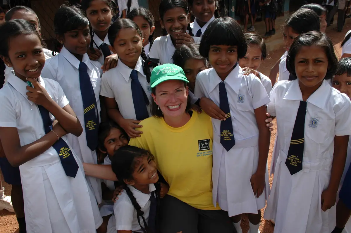 Volunteer Maureen Quiery, in a green cap and a yellow T-shirt, kneels smiling among schoolgirls in white uniforms and navy ties in Sri Lanka in 2011.