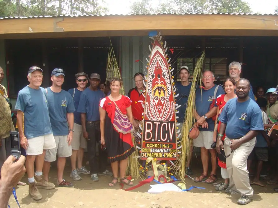 A volunteer team and community members stand beside a carved, painted sign reading "BTCV School No. 3, Brikiti Elementary School, Maprik, PNG" in Papua New Guinea.