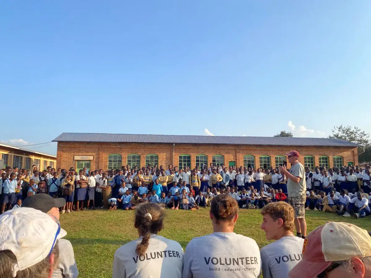 A large group of students in uniform are gathered outdoors on a field, listening to an adult male speaker in front of a brick building. The backs of several people wearing "VOLUNTEER" shirts are visible in the foreground.