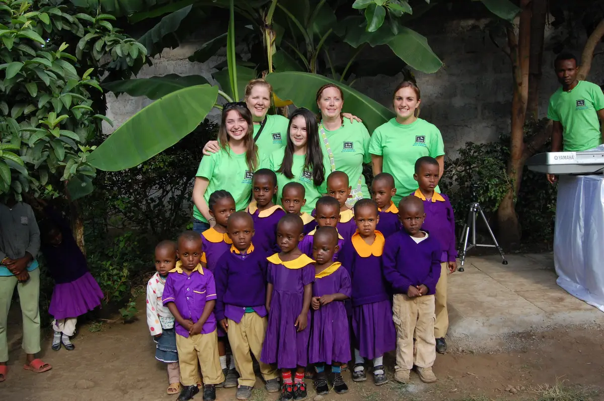 Five volunteers in green T-shirts stand behind a group of young students in purple and yellow uniforms in a garden in Tanzania.