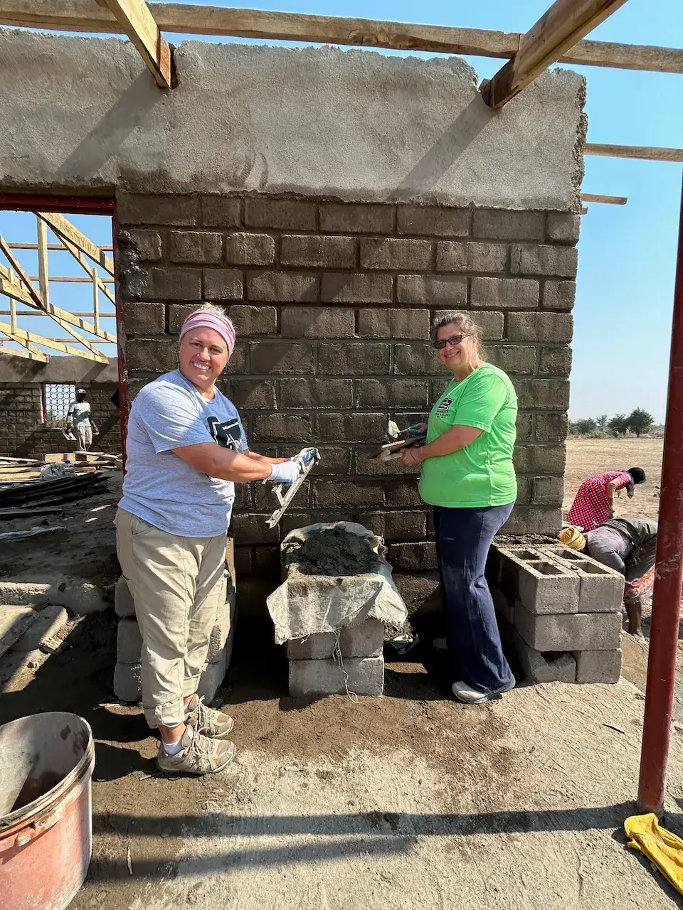Two women are building a block wall, applying mortar in a sunny outdoor construction setting.