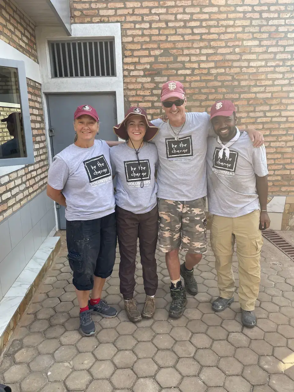 Four volunteers wearing matching gray t-shirts and red baseball caps pose in front of a brick building.