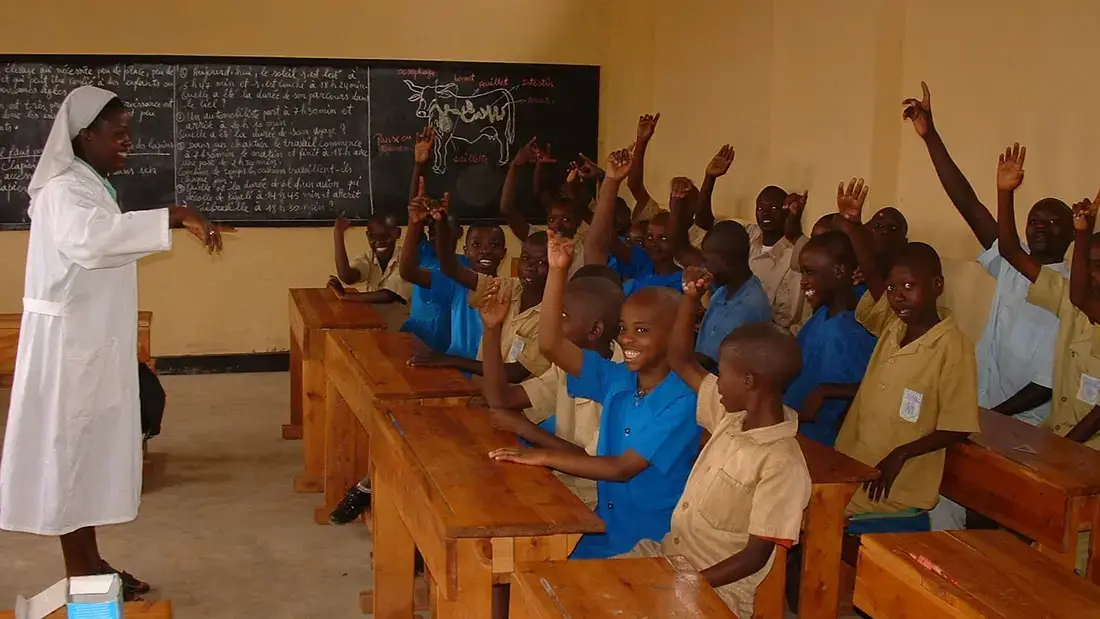 Students in blue and khaki uniforms raise their hands at wooden desks while a teacher in a white habit leads a lesson at the chalkboard of a new classroom in Rwanda.