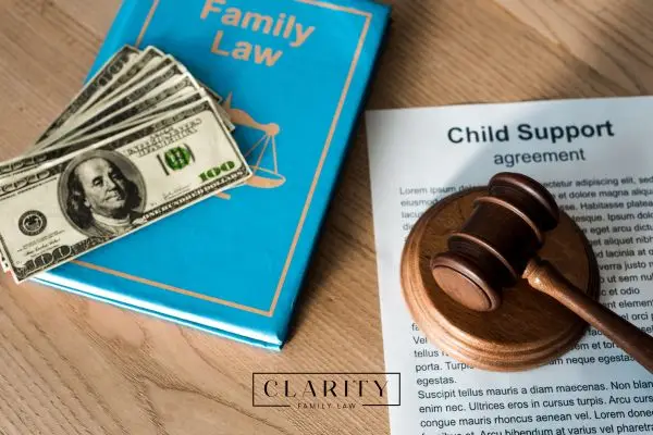 A family law book, a stack of hundred-dollar bills, a child support agreement, and a judge’s gavel on a wooden table.