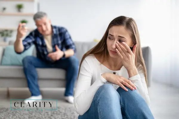 A woman sits on the floor, crying and wiping her eye, while a man in the background gestures angrily.