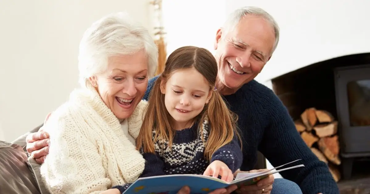 A smiling grandmother and grandfather sitting closely with their granddaughter, reading a book together