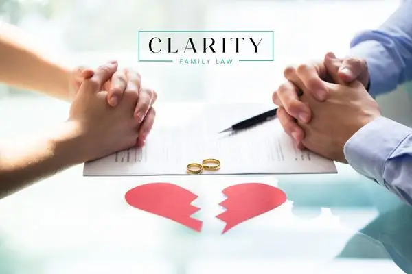 hands clasped over a document featuring two wedding rings and a pen, with a paper cutout of a broken heart