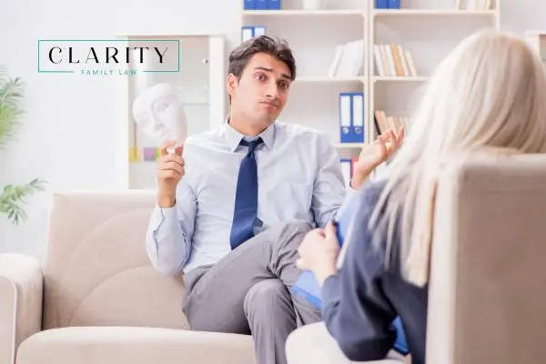 A man sits across from a therapist, holding a white mask and shrugging.