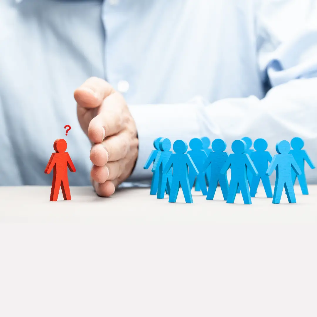 Only the torso and arm of a man wearing a button down shirt are visible. His hand is positioned on a desktop vertically, with his pinky finger on the bottom, pressed into the desktop and his thumb at the top. His hand is intentionally forming a barrier between a group of people (represented as little blue 3D cutouts standing around in a group) and a single person (shaped as a little red 3D cutout standing by itself). The red person has a small red question mark above their head as if they can't understand why the man is excluding them from the group. The image symbolizes the uncertainty that some people feel about whether discrimination is the root cause of their workplace situation and whether they need to speak with a discrimination lawyer.