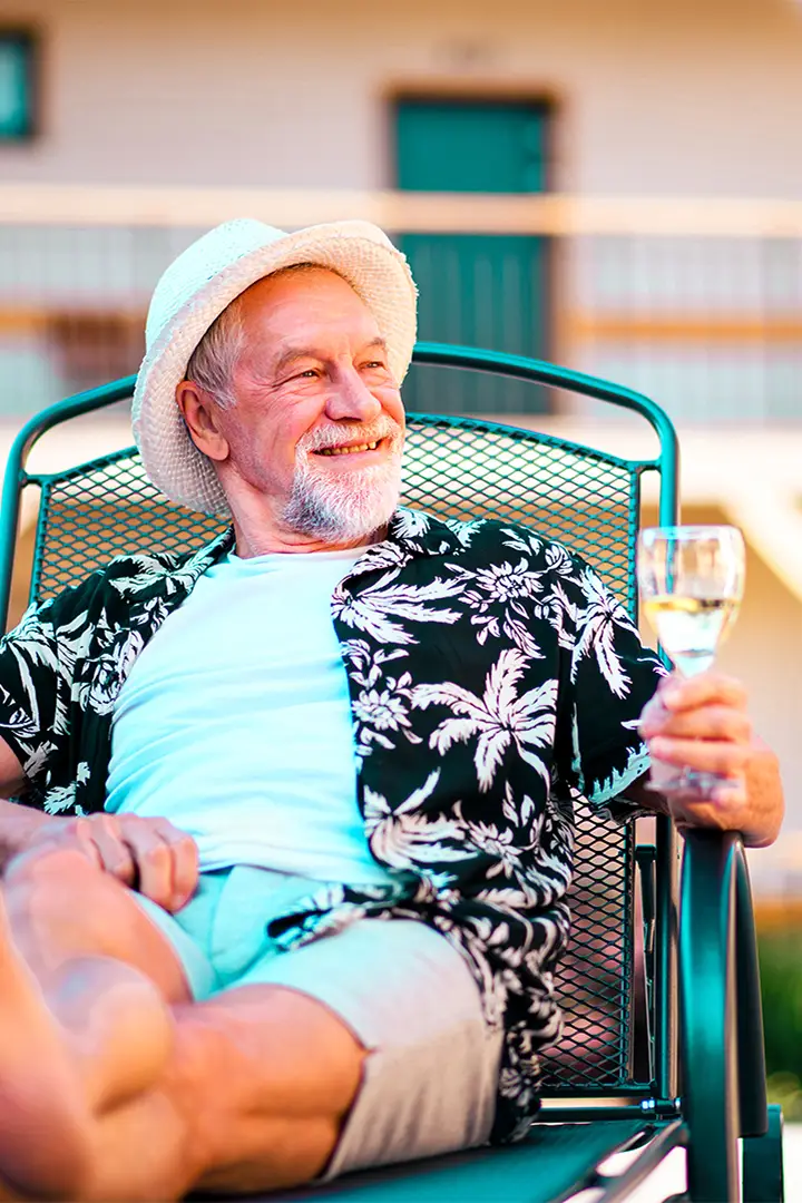 Smiling older man in a straw hat and palm-print shirt relaxing outdoors with a glass of wine