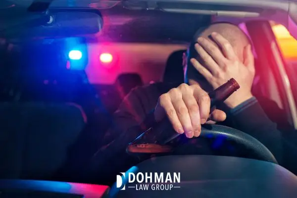 Driver pulled over by police, holding a bottle, with their hand over their face; police lights flashing.
