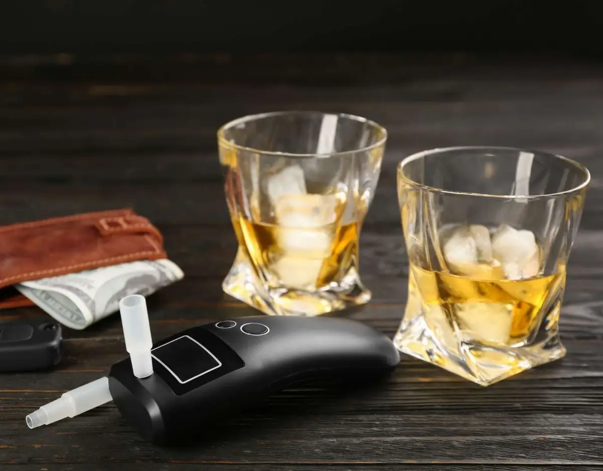 A breathalyzer on a wooden table next to two glasses of whiskey, a car key, and a wallet.