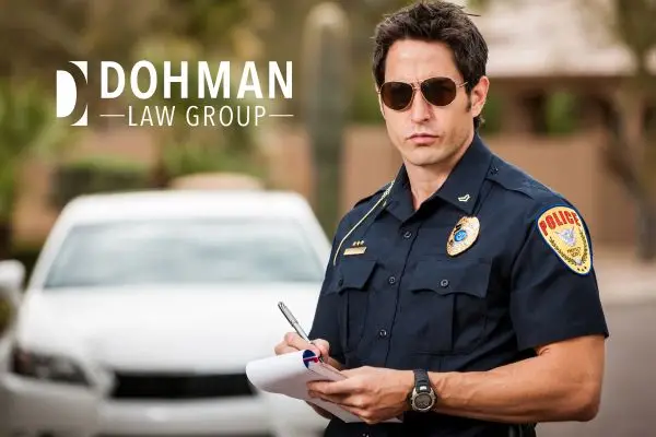A male police officer in sunglasses writing on a notepad in front of a white car.
