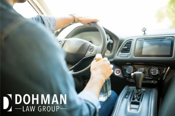 A person is driving a car while holding an open bottle of clear liquid.