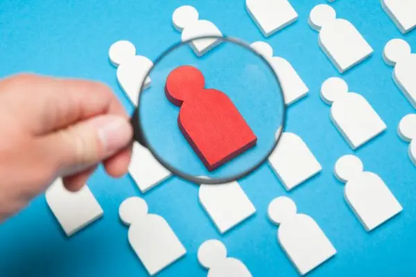 A magnifying glass over a red person icon in a crowd of white person icons, on a blue background.
