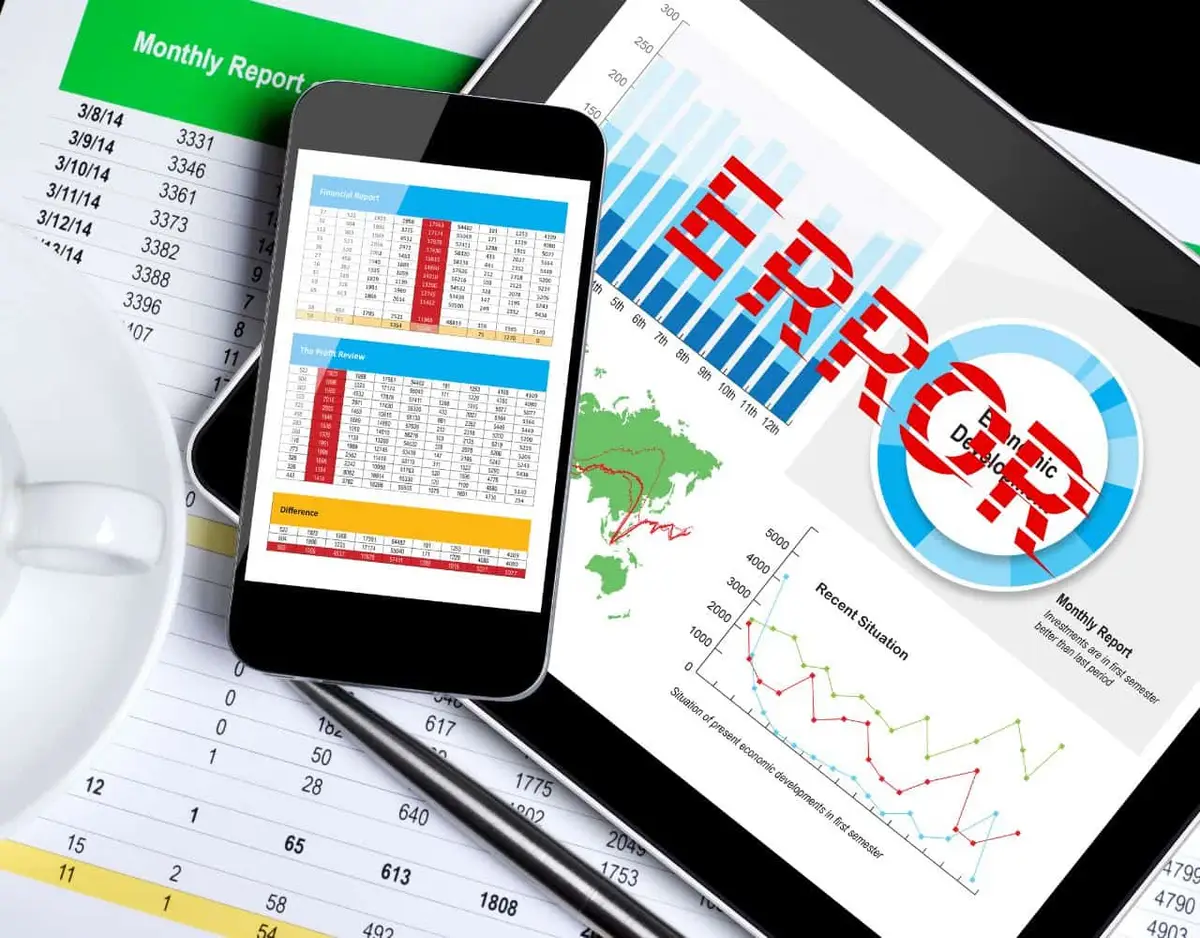 A tablet and smartphone display financial reports, graphs, and the word "ERROR" in red. Papers with numbers and calculations are in the foreground, with a white cup in the top left.