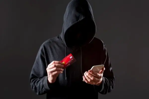 A hooded figure holds a red credit card and a smartphone, their face obscured