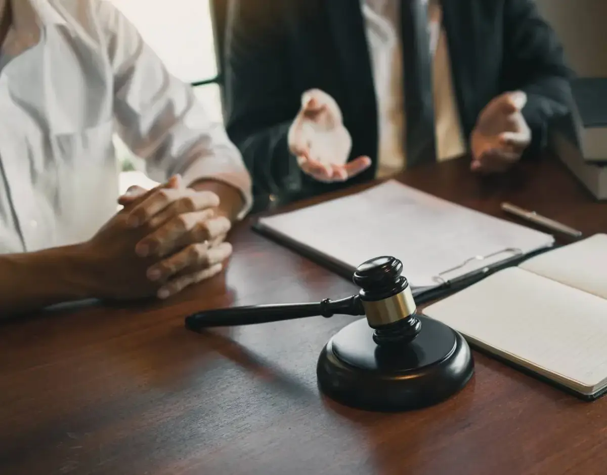 Close-up of a judge's gavel on a wooden desk with a defense attorney and client blurred in the background.