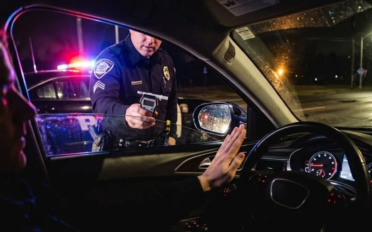 Driver raising a hand to refuse a Breathalyzer held by a police officer outside a car window at night.
