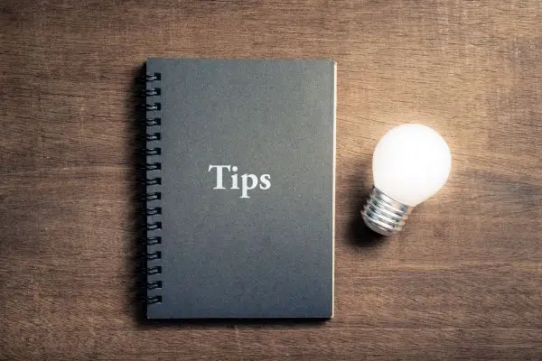 A black spiral notebook with the word "Tips" on the cover next to a glowing lightbulb on a wooden table.