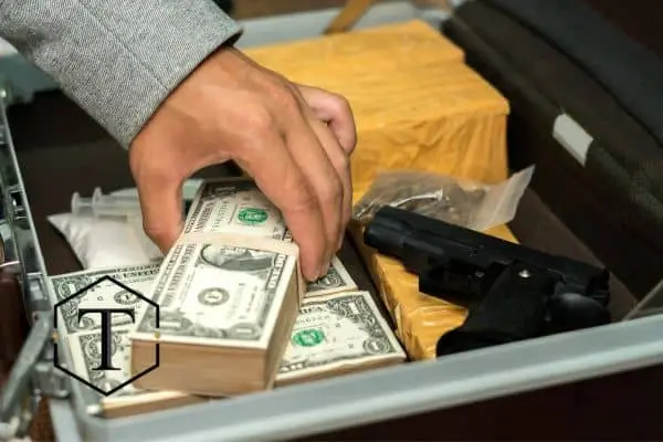Hand reaching for stacks of money next to a gun and drugs in a briefcase.